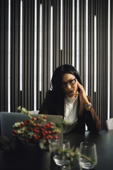 A businesswoman is seated at a desk in a contemporary office, deep in thought, with stylish vertical lighting casting soft shadows. She is dressed in professional attire and appears focused