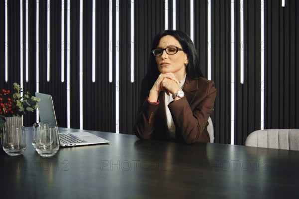 A thoughtful woman with glasses sits at a modern office desk. She has long dark hair and is dressed in a brown suit