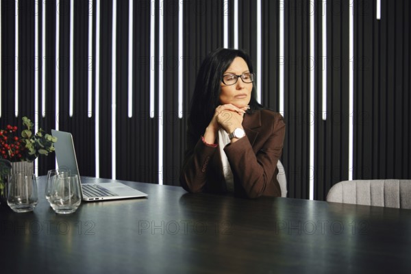 A professional woman sits at a modern desk in an elegant office space. She rests her chin on her hands, lost in contemplation with a laptop open nearby. Soft lighting creates a reflective atmosphere