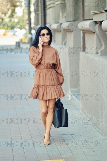 A fashionable woman strolls down the city sidewalk wearing a brown dress and sunglasses, carrying a black handbag. Warm sunlight illuminates the bustling urban surroundings
