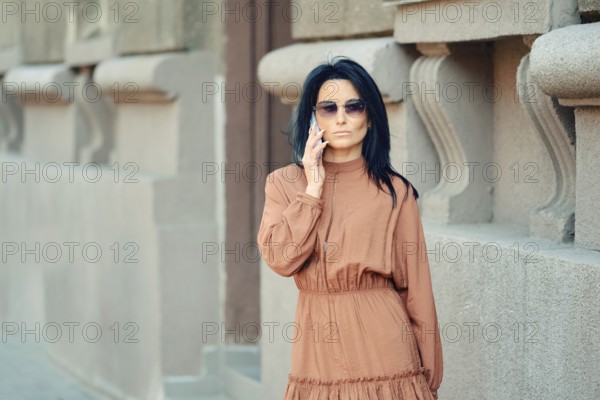 A woman wearing a brown dress and sunglasses engages in a phone conversation while passing by the building