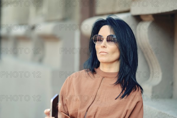 A woman wearing stylish sunglasses stands next to a historic building, casually holding her smartphone