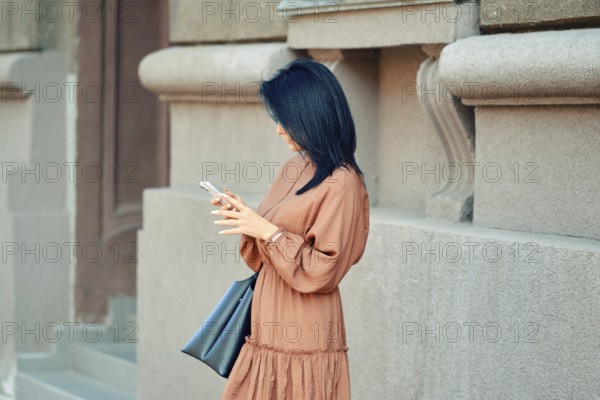 A woman in a flowing brown dress and a handbag stands against a stylish urban building. She is ordering a taxi online using the app in her smartphone