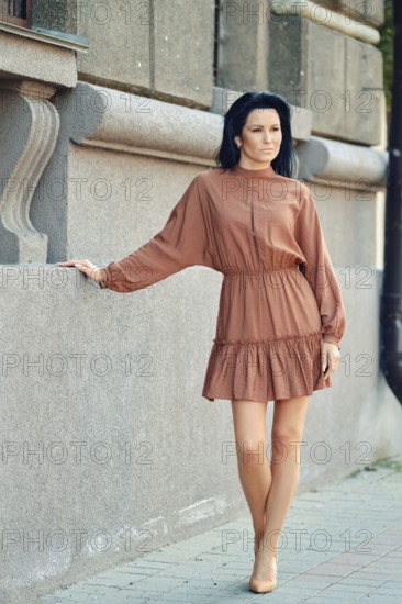 A relaxed woman stands by a textured stone wall, wearing a stylish brown dress with a flowing skirt. Sunlight highlights her elegant look as she enjoys a pleasant day outdoors in the city