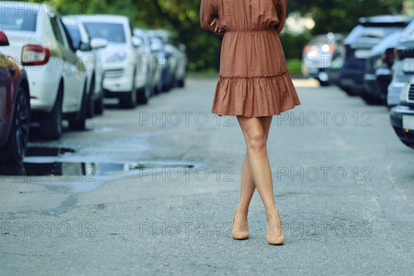 Unrecognizable woman is walking down a city street, wearing a brown dress and high heels. The pathway is lined with parked cars, reflecting a vibrant urban setting on a sunny day