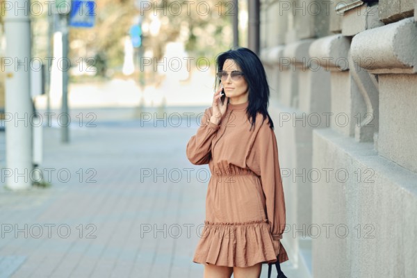 A fashionable woman dressed in a brown dress engages in a phone conversation while standing on a busy city sidewalk during the day. She enjoys the warm sunlight and urban atmosphere