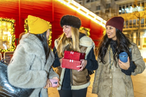 Three cheerful young women holding christmas gifts and enjoying a winter night out in a decorated city
