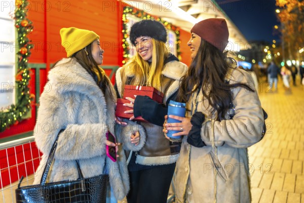Three cheerful women holding christmas gifts and hot drinks, enjoying christmas shopping at a decorated night market
