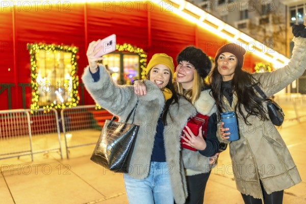 Three happy women taking selfie with smartphone in front of illuminated christmas market kiosk