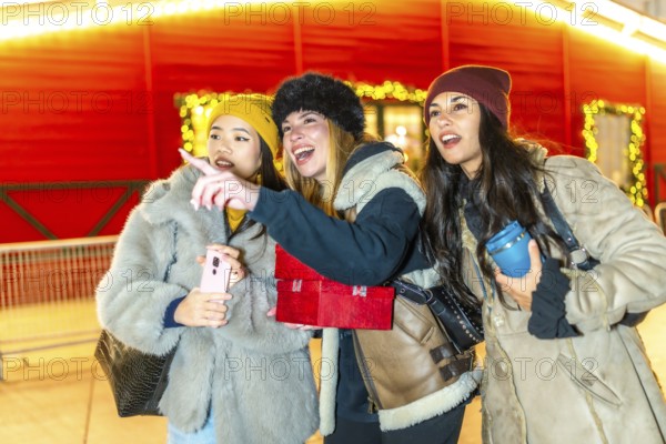 Three young women holding christmas gifts and a smartphone are enjoying the christmas spirit, pointing at something interesting in a brightly lit market stall