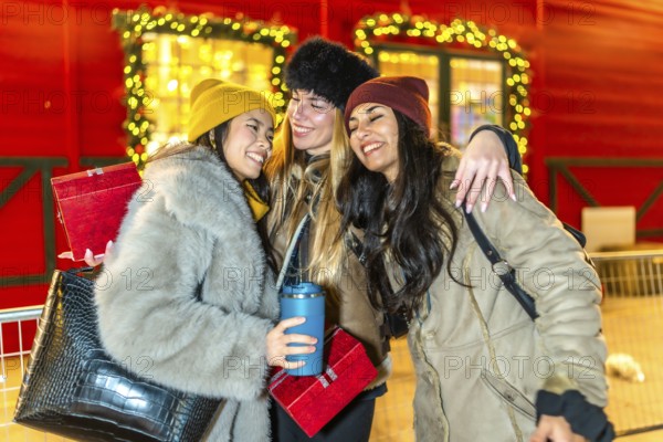 Three happy female friends hugging at a decorated christmas market stall at night, holding christmas presents and a reusable cup