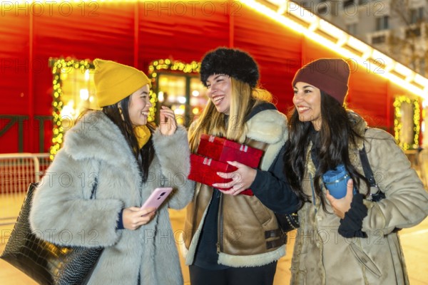 Three cheerful young women holding christmas gifts, enjoying christmas market at night, winter holidays shopping concept