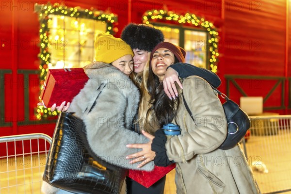 Three happy female friends are hugging at a christmas market at night, enjoying the festive atmosphere and holding gifts