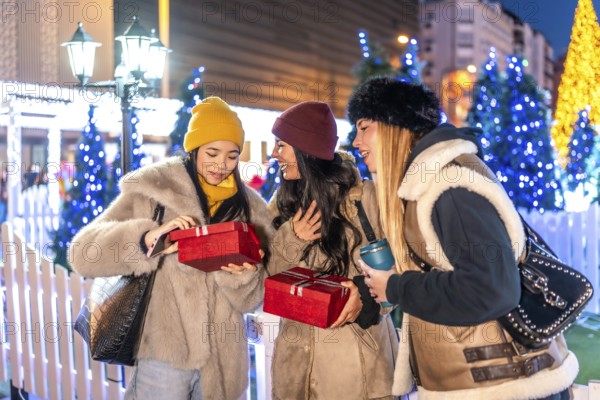 Cheerful young women exchanging christmas gifts at a decorated christmas market in the city at night