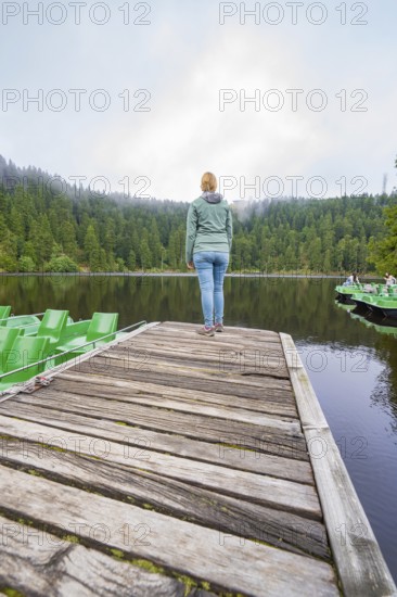 Woman standing alone on a wooden jetty in front of a quiet lake surrounded by forest, Mummelsee, Seebach, Ortenau District, Black Forest, Germany