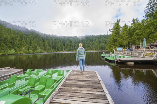 Woman on a jetty at the lake, surrounded by forest and boats under a cloudy sky, Mummelsee, Seebach, Ortenau District, Black Forest, Germany