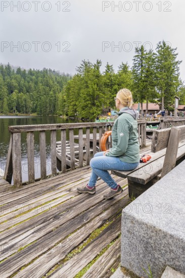 A woman sits relaxed on a wooden bench overlooking the tranquil lake and forest, Mummelsee, Seebach, Ortenau District, Black Forest, Germany