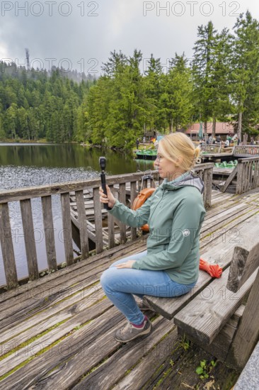 Woman sitting on a wooden footbridge and holding a camera over a lake, Mummelsee, Seebach, Ortenau district, Black Forest, Germany