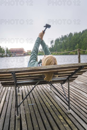 Woman lying on a bench by the lake and holding a camera, Mummelsee, Seebach, Ortenau district, Black Forest, Germany