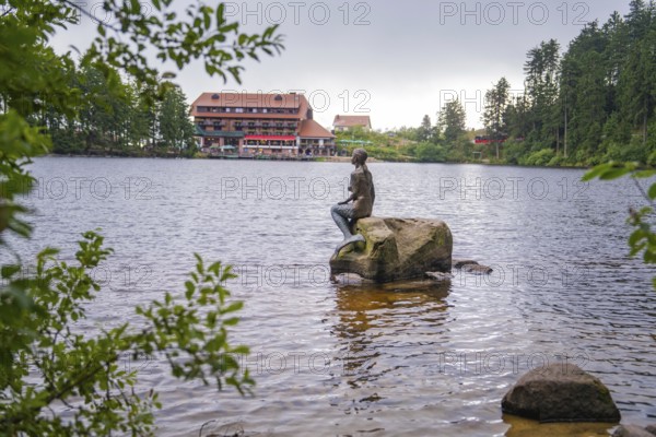 A statue sits on a rock in the lake, with a building in the background, Mummelsee, Seebach, Ortenau district, Black Forest, Germany