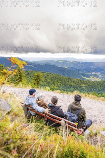 Three people on a bench enjoy the wide view over wooded mountains and valleys, Mummelsee, Seebach, Ortenau district, Black Forest, Germany