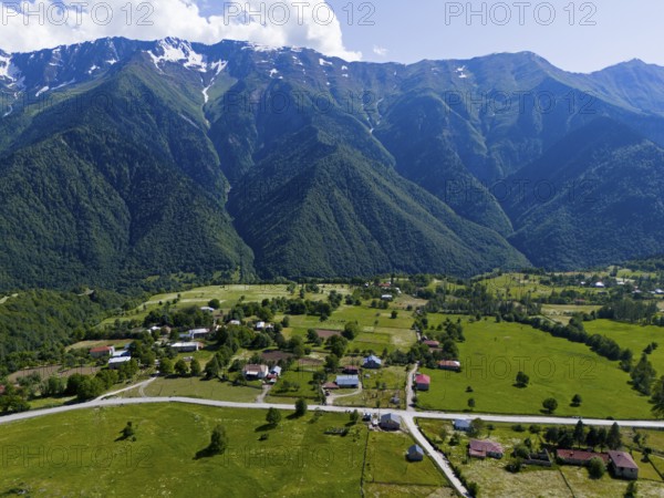 A small village in the middle of a vast green landscape with high mountains in the background, aerial view, landscape near Iskari, Mingrelia and Upper Svaneti, Samegrelo-Zemo Svaneti, Svaneti, High or Greater Caucasus, Georgia