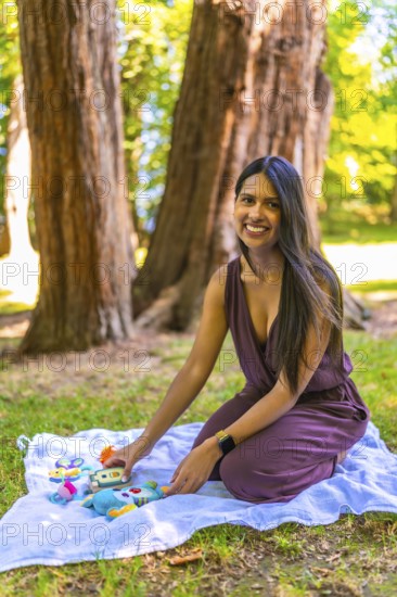 Latin mother arranging colorful baby toys on a blanket in a park, enjoying a sunny day outdoors