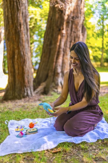 Happy young latin mother kneeling on a blanket in a park, preparing vibrant baby toys while enjoying the sunny outdoor atmosphere