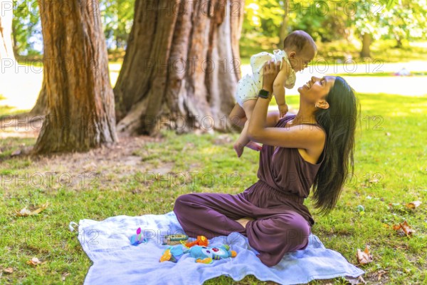 Young latin mother playing with her baby girl in a park, lifting her in the air and smiling, sitting on a blanket