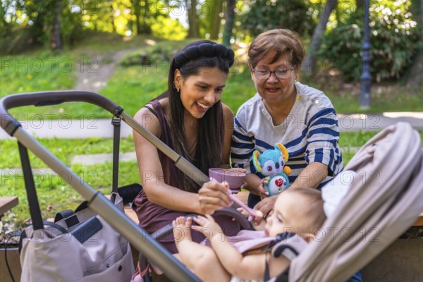 Grandmother, mother and baby girl enjoying time together at the park, feeding the baby girl and playing with a plush toy