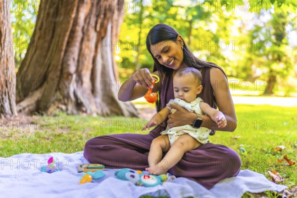 Latin mother holding her baby daughter and playing with colorful toys while sitting on a blanket in a park