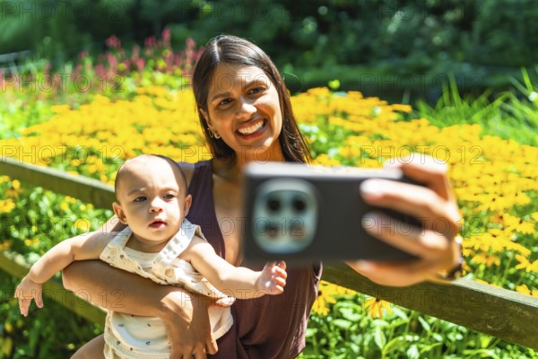 Happy latin mother holding her baby daughter, taking a selfie with a smartphone in a garden full of yellow flowers