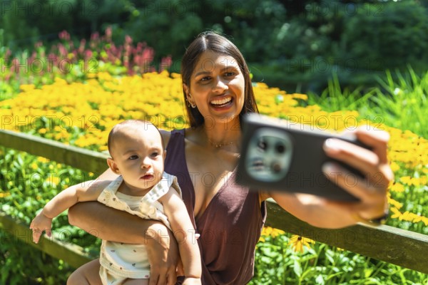 Happy latin mother holding her baby daughter and taking a selfie with smartphone in a beautiful garden full of yellow flowers