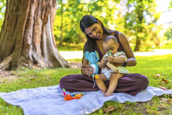 Latin mother and her baby daughter enjoying playtime with colorful toys while sitting on a blanket in a park