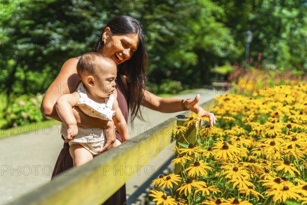 Latin mother holding her baby daughter and showing her yellow flowers in a park during a sunny day
