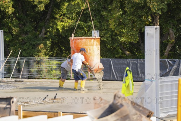Construction workers pouring concrete from a concrete mixer on a building site with lattice fence and trees in the background, car park construction, Calw, Germany