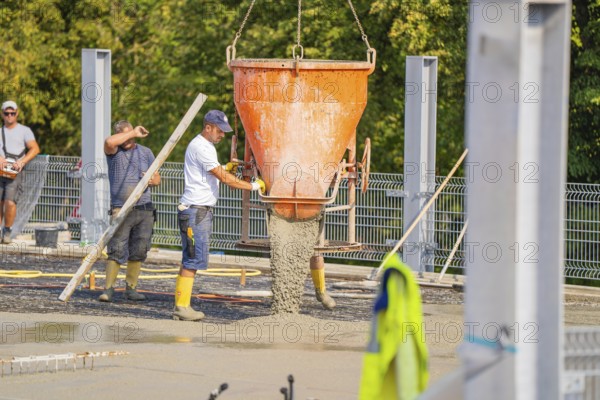 Two workers pouring concrete from large mixer tubs, surrounded by safety measures and mesh fencing, car park construction, Calw, Germany
