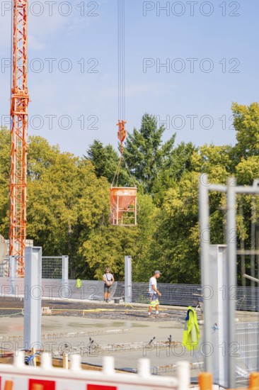 Crane lifts a concrete bucket over an almost finished building site. Two construction workers, surrounded by green foliage and a clear sky, car park construction, Calw, Germany