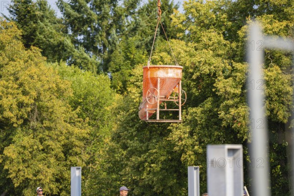 An orange-coloured concrete bucket hovers over a building site. Trees and a clear sky in the background, car park construction, Calw, Germany