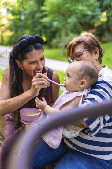 Mother feeding her baby a meal in the park, with grandmother holding the child, enjoying family time together