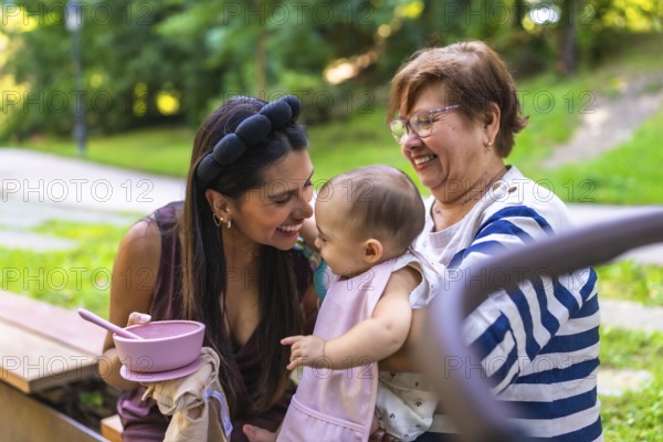 Grandmother holding her granddaughter while mother is feeding her in a park, enjoying family time together