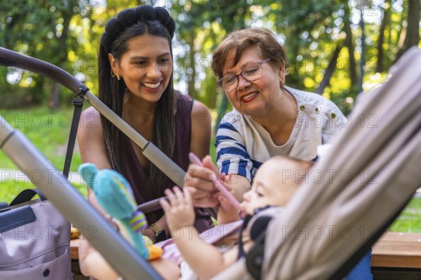 Grandmother lovingly feeding baby girl with a spoon while mother watches from the stroller in a sunny park filled with green trees