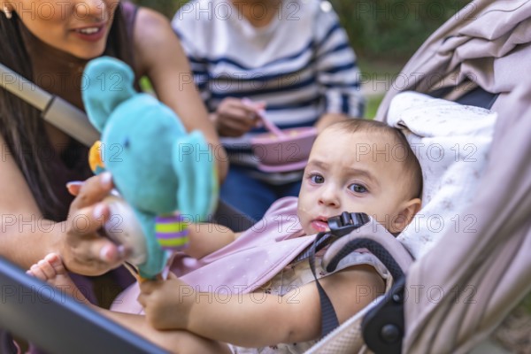 Mother showing a stuffed toy to her baby daughter sitting in a stroller, while grandmother is eating in the background, enjoying a sunny day outdoors