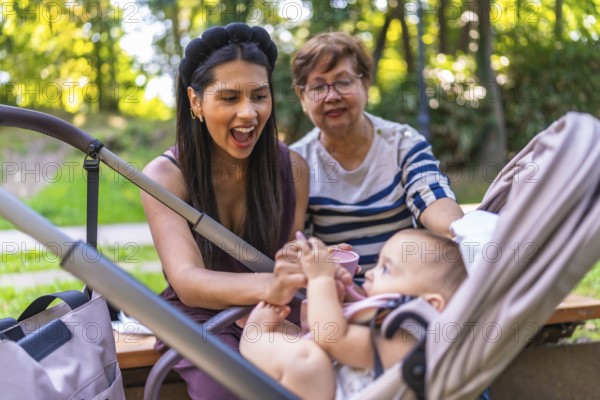 Grandmother, mother and baby girl enjoying a sunny day at the park, feeding the baby sitting in a stroller