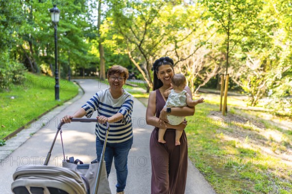 Happy latin family walking in a park, grandmother pushing a stroller and mother holding her baby