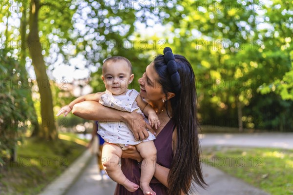 Happy latin mother smiling while holding her baby girl in a park, enjoying a sunny summer day filled with warmth and joy