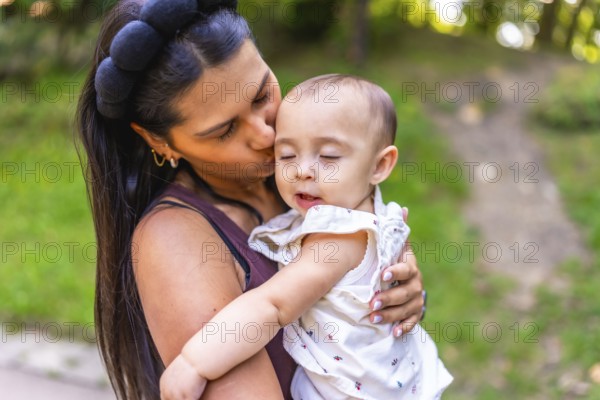 Young latin mother kissing her baby daughter while holding her lovingly in her arms, surrounded by the beauty of a green park