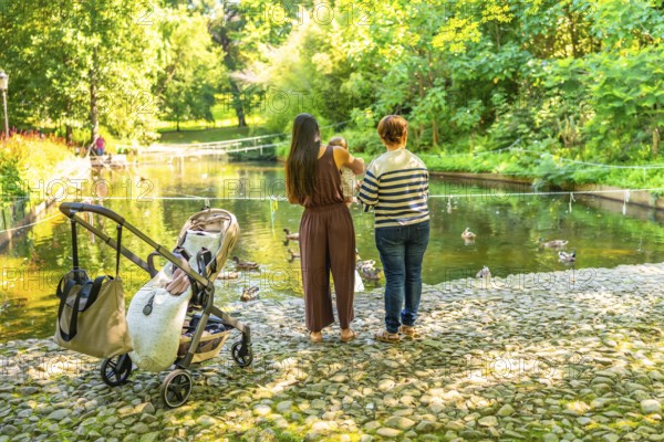 Mother, grandmother and baby girl enjoying a sunny day by the pond, watching ducks swim in a park