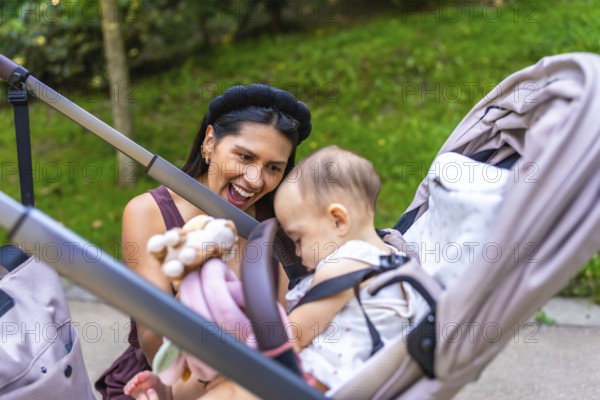 Happy latin mother showing toys to her baby daughter sitting in a stroller, enjoying a sunny day together in a park