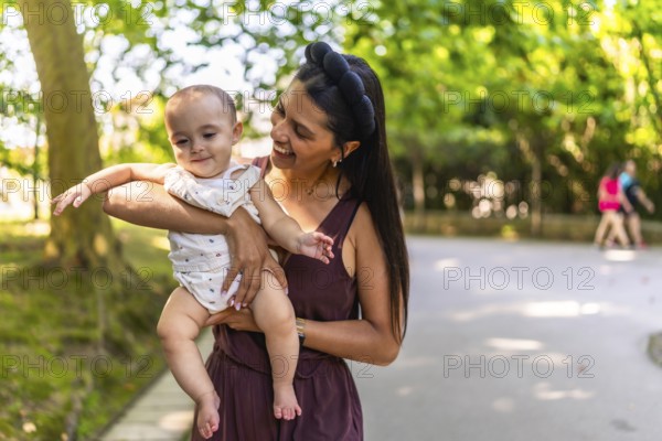 Happy latin mother holding and playing with her baby daughter in a park, enjoying a sunny day outdoors